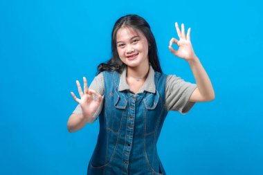 Gülümseyen genç Asyalı kadın kendine güvenle el hareketlerini yapıyor, kot elbise ve gri gömlek giyiyor, stüdyo ışıklarında parlak mavi bir arka plana karşı duruyor..