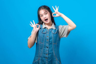 Heyecanlı, uzun saçlı genç Asyalı kadın gülümseyen ve parlak mavi bir arka plana karşı anlamlı bir şekilde poz veren iki el işareti yapıyor. Günlük kot ve gri elbise giyiyor..