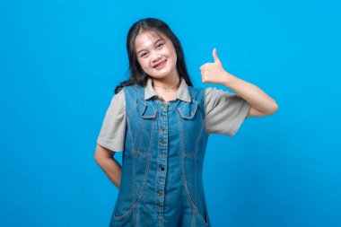 Gülümseyen Asyalı genç bir kadının portresi, başarı için onay, güven ve pozitif vücut dilini gösteren mavi bir arkaplan önünde dikilirken başparmağını kaldırıyor..
