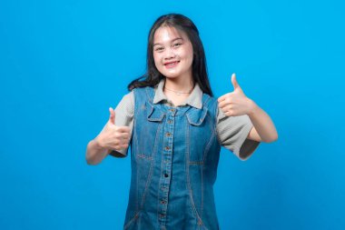 Neşeli genç Asyalı kadın, parlak bir gülümseme ile iki baş parmak kaldırıyor, mavi bir arka plan karşısında duruyor, onayı, başarıyı, memnuniyeti ve olumlu geri bildirimi temsil ediyor..