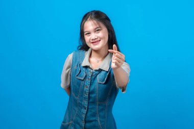 Gülümseyen Asyalı genç bir kadın, Koreli bir kalp hareketiyle, kot elbise ve gri gömlek giyerek, parlak mavi bir arka plana karşı duran, stüdyo portresi.