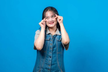 Mutlu genç Asyalı kadın parmaklarıyla sevimli küçük bir kalp hareketi yapıyor, tatlı tatlı gülümsüyor, rahat bir kot elbise giyiyor, parlak mavi arka planda izole edilmiş..