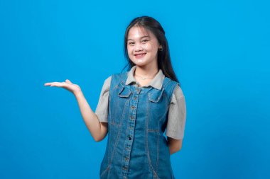 Gülümseyen Asyalı genç bir kadın, açık avuçlarıyla sunum yaparken, koyu mavi arka planda duruyor, kot elbise ve günlük gömlek giyiyor. Reklam ve tanıtım için mükemmel..