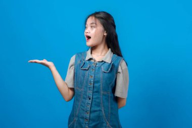 Şaşırmış genç Asyalı kadın yan bakıyor ve açık avucunda bir şey sunuyor, kot elbise ve gri gömlek giyiyor, parlak mavi stüdyo arka planında güvenle duruyor.