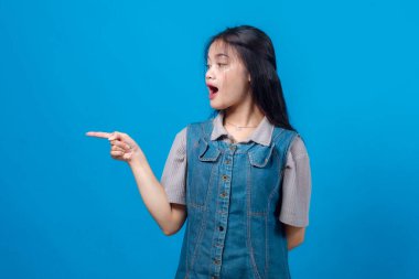 Asyalı genç bir kadın ağzı açık, parlak mavi bir arka planda kot elbise giyiyor. Reklam, afiş veya promosyon tasarımı için stüdyo çekimi.