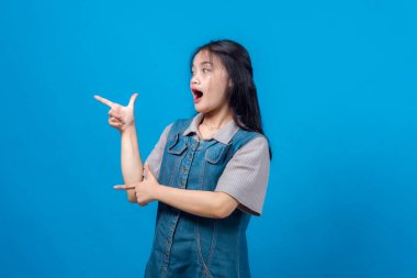 Şaşırmış genç Asyalı kadın parmak tabancası hareketiyle yanlamasına işaret ediyor, ağzı açık kalmış. Parlak mavi arka planda stüdyo portresi. Eğlenceli, anlamlı ve çekici..