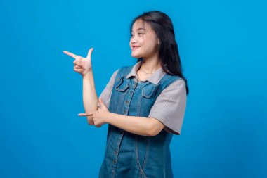 Gülümseyen Asyalı genç bir kadın parmak tabancası hareketi yapıyor ve bir tarafı işaret ediyor, parlak mavi arka plana karşı duruyor. Neşeli ifade, stüdyo çekimi, promosyon konsepti veya reklam..