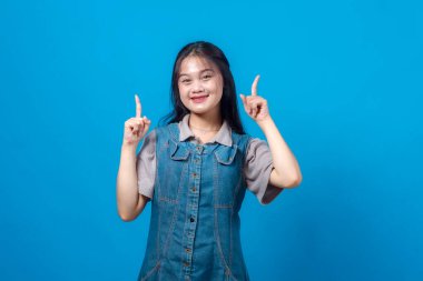Kot elbiseli mutlu genç Asyalı kadın her iki işaret parmağını yukarı kaldırarak güvenle gülümsüyor, düz mavi bir arkaplan üzerinde duruyor, reklam için ideal ya da kopyalama alanı için.