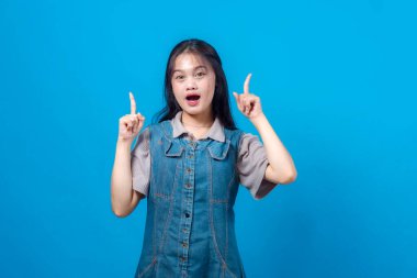 Kot elbiseli heyecanlı genç Asyalı kadın her iki işaret parmağıyla yukarıyı gösterirken gülümsüyor ve şaşırmış görünüyor, canlı mavi bir stüdyo geçmişine karşı duruyor, izole edilmiş..