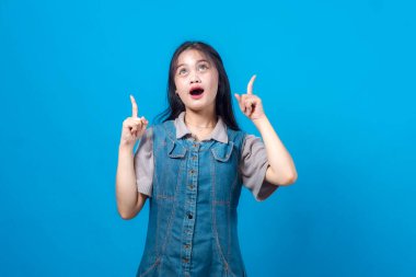 Kot elbiseli genç Asyalı kadın her iki işaret parmağıyla yukarıya bakıyor ve hayret verici bir ifadeyle bakıyor, mavi arka planda kopya alanı ya da reklamlar için izole edilmiş..