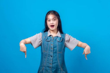 İki eliyle aşağıya bakan genç Asyalı kadın, şaşırmış yüz ifadesi, mavi arka planda izole edilmiş. Promosyon veya duyuru konsepti.