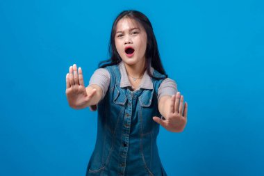 Ciddi bir ifadeyle genç Asyalı kadın dur işareti olarak iki elini de kaldırdı. Mavi arka planda izole edilmiş. Uyarı, reddetme, protesto etme veya sınır koyma kavramı.