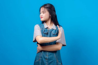 Asyalı bir kadın onaylamayan bir ifadeyle başka tarafa bakıyor, bir kolunu kaldırıyor ve diğer kolunu çaprazlıyor. Mavi arka planda izole edilmiş. Reddedilme ve onaylanmama kavramı.