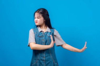 Asyalı genç bir kadın yüzünü buruşturuyor ve bir eliyle sert bir dur hareketi yapıyor, diğer eliyle diğer kolunu çapraz yapıyor, güçlü bir şekilde reddediyor ya da protesto ediyor. Mavi arka planda izole edilmiş, stüdyo çekimi..