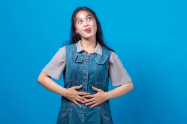 Neşeli, genç Asyalı bir kadın iki eliyle karnını tutarken şakacı bir şekilde dilini uzatıyor. Eğlenceli ve tasasız bir yüz ifadesi sergiliyor. Mavi arkaplan ile stüdyo portresi.