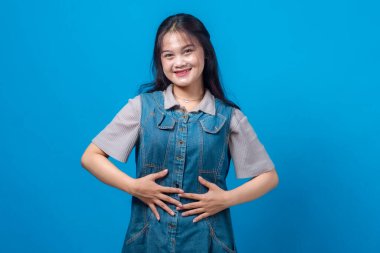 Genç Asyalı kadın iki elini karnına koyup güvenle gülümsüyor, mutluluğu, tatmini ya da sindirim sağlığını ifade ediyor. Mavi arkaplanda stüdyo resmi, ön görünüm.