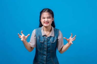 Asyalı genç bir kadın elleri pençe gibi havada sinirli bir yüz ifadesi takınıyor, hayal kırıklığı ya da öfke gösteriyor, mavi arka planda soyutlanmış. Duygu, tutum ve ifade.