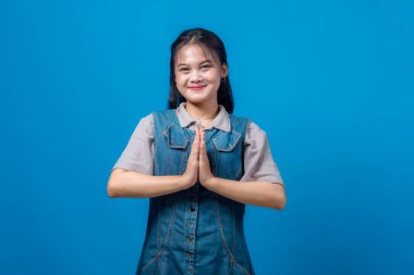 Genç Asyalı kadın gülümsüyor ve avuçlarıyla geleneksel bir karşılama hareketi yapıyor. Saygı, minnettarlık ve barışı simgeliyor. Mavi arkaplanda stüdyo resmi.