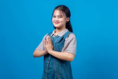 Genç Asyalı kadın gülümsüyor ve avuçlarıyla geleneksel bir karşılama hareketi yapıyor. Saygı, minnettarlık ve barışı simgeliyor. Mavi arkaplanda stüdyo resmi.
