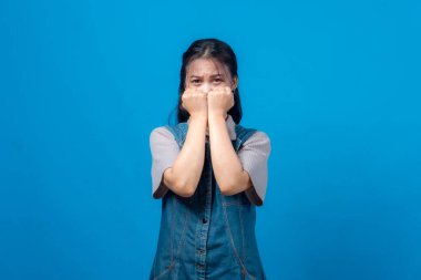 Genç Asyalı kadın portresi korku ya da endişeyi yüzüne yakın yumruklarıyla dışa doğru bakıyor. Mavi arka planda stüdyo çekimi, duygusal tepki, kot elbise..
