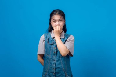 Genç Asyalı kadın portresi korku ya da endişeyi yüzüne yakın yumruklarıyla dışa doğru bakıyor. Mavi arka planda stüdyo çekimi, duygusal tepki, kot elbise..