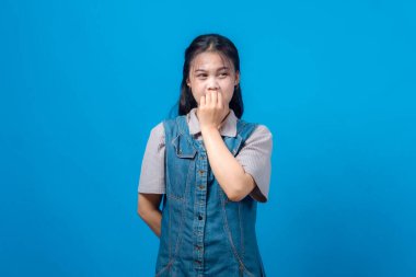 Genç Asyalı kadın portresi korku ya da endişeyi yüzüne yakın yumruklarıyla dışa doğru bakıyor. Mavi arka planda stüdyo çekimi, duygusal tepki, kot elbise..