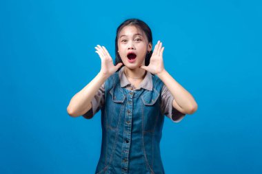 Asyalı genç bir kadının portresi, elleri ağıza yakın bağırıyor, sürpriz ya da heyecan ifade ediyor, mavi arka planda izole edilmiş. Duyuru veya uyarı kavramı.