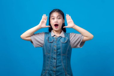 Heyecanlı genç Asyalı kadın, ağzı açık ve elleri havada, şok olmuş ya da şaşkın bir ifade sergiliyor, mavi bir arkaplanın önünde duruyor, şaşırmış görünüyor..