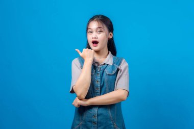 Şaşırmış genç bir Asyalı kadının portresi başparmağı yana, ağzı açık ve gözleri açık, mavi bir arkaplanın önünde duruyor. Tanıtım konsepti.