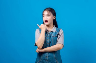 Şaşırmış bir ifadeye sahip genç bir Asyalı kadın, canlı mavi bir arkaplan önünde kendinden emin bir şekilde başparmağını yana çeviriyor..