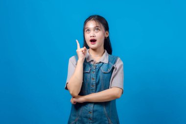 Şaşırmış yüzlü genç bir Asyalı kadının portresi, işaret parmağıyla yukarıya doğru, sanki parlak bir fikri varmış gibi, sağlam mavi bir arka planda duruyor..