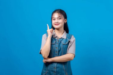 Neşeli genç Asyalı kadın işaret parmağını kaldırırken gülümsüyor, bir fikir ya da çözüm öneriyor, özgüvenle mavi arka planda duruyor..