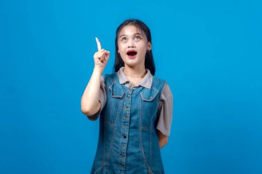 Heyecanlı genç Asyalı kadın işaret parmağını yukarı kaldırıp sanki parlak bir fikir edinmiş gibi neşeli bir ifadeyle sade bir mavi arkaplanın önünde duruyor..