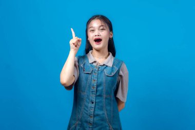 Neşeli, genç Asyalı bir kadın heyecanlı bir ifadeyle yukarı işaret ediyor, ağzı açık, sıradan bir kot elbise giyiyor, stüdyo çekiminde düz mavi arka plana karşı duruyor..