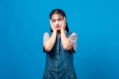 Genç Asyalı kadın stresli ve hayal kırıklığına uğramış görünüyor, iki eliyle yanaklarını bastırıyor, kot elbise giyiyor, endişeli bir ifadeyle stüdyoda sade mavi arka planda duruyor..