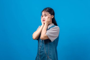 Genç Asyalı kadın stresli ve hayal kırıklığına uğramış görünüyor, iki eliyle yanaklarını bastırıyor, kot elbise giyiyor, endişeli bir ifadeyle stüdyoda sade mavi arka planda duruyor..