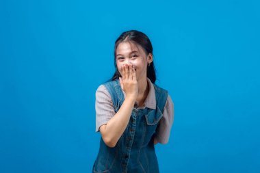 Genç Asyalı kadın utangaç gülümseyerek ağzını kapatıyor, utancını ya da kahkahasını ifade ediyor, günlük kıyafetler giyiyor, neşeli bir stüdyo çekiminde mavi arka planda duruyor..