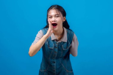 Genç Asyalı kadın bağırıyor ya da eli ağzının kenarında bağırıyor, aciliyet ya da duyuru yapıyor. Canlı mavi arkaplanı olan bir stüdyo portresi, reklam kavramları için mükemmel..