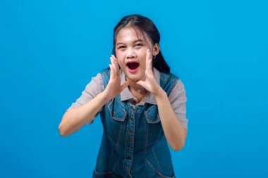 Genç Asyalı kadın iki eliyle bağırıyor, ağzını çerçeveliyor, heyecanını dile getiriyor ya da yüksek sesle bağırıyor. Parlak mavi arkaplanlı stüdyo çekimi, pazarlama ve reklam kavramları için ideal.