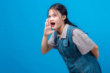 Genç Asyalı bir kadın bağırıyor ya da ağızdan ağıza sesleniyor, mavi arka planda duruyor. Etkileyici, enerjik ve hevesli görünüyor..