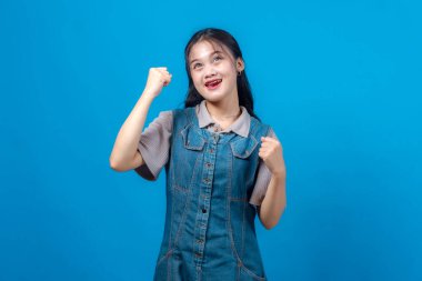 Kot elbiseli neşeli genç Asyalı kadın yumruklarını kaldırıyor ve heyecanla yukarı bakıyor, pozitifliğini, hırsını ve umudunu açık mavi stüdyo geçmişine karşı gösteriyor..