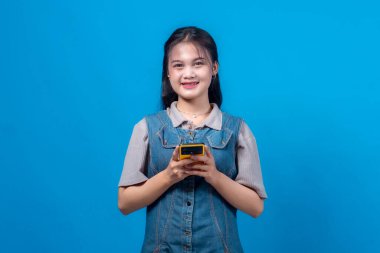 Çekici genç bir kadın akıllı bir telefon tutuyor, parlak mavi bir arka plan karşısında kameraya samimi bir şekilde gülümsüyor. Kendine güveni ve dostça ifadesi bu resmi modern reklamcılık, sergi teknolojisi, dijital iletişim ve varsayım için ideal kılıyor.