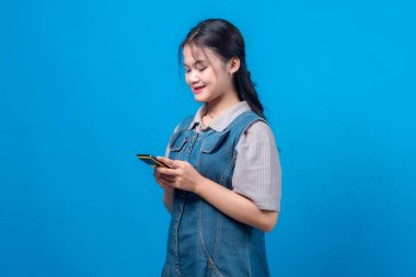 Akıllı telefonuna odaklanmış genç bir kadın, parlak mavi bir arka planda ince bir gülümseme ile. Bu modern portre küresel reklam kampanyaları, vitrin bağlantısı, dijital bağlantı, mobil uygulama kullanımı ve çevrimiçi etkileşim için idealdir.