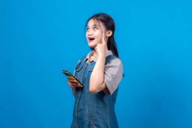 Akıllı telefon tutan düşünceli genç bir kadın parmağını şakağına koyup yukarı bakar. Mavi bir arka plana karşı bir fikir veya düşünce ileri sürer. Bu ilgi çekici portre küresel reklamcılık, problem çözme, ilham vermek için mükemmel.