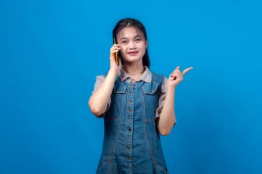 Genç Asyalı bir kadın akıllı telefonda konuşurken gülümsüyor ve yan tarafı işaret ediyor, kot bir elbise giyiyor, mavi arka planda izole edilmiş. Müjde kavramı veya yararlı öğüt.