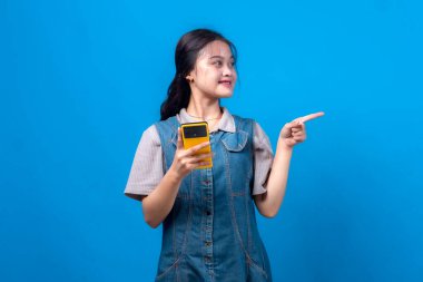 Sarı bir akıllı telefon tutan neşeli genç Asyalı kadın gülümserken yan tarafı işaret ediyor mavi arka planda izole edilmiş sıradan bir kot giysisi giyiyor. Dijital reklamlar için kavram.