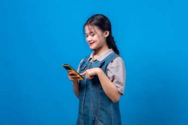 Sarı bir akıllı telefon kullanırken gülümseyen Asyalı genç bir kadının yandan görünüşü. Kot pantolon giymiş, parlak mavi bir arka planda izole edilmiş, rahat ve mutlu bir ruh hali sergiliyor..