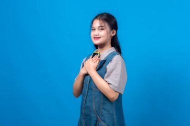 Gülümserken göğsüne yakın sarı bir akıllı telefon tutan genç bir Asyalı kadının portresi. Sıradan bir kot giysisi giyiyor. Fotokopi çekilen mavi arka planda soyutlanmış..