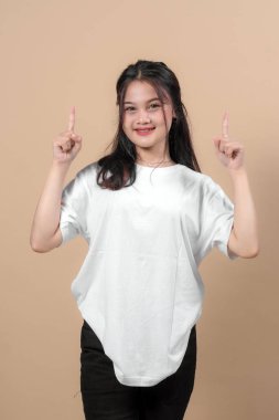 Beyaz tişörtlü genç Asyalı kadın gülümseyip işaret parmaklarını yukarı kaldırırken bej arka planda duruyordu. Reklam ve tanıtım içeriği için ideal.
