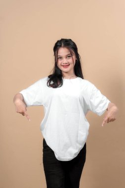 Beyaz tişörtlü, gülümseyen ve iki eliyle işaret eden genç Asyalı kadın, bej bir arka plana karşı duruyor, reklam ya da taklit tasarım için ideal..